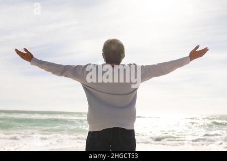 Vista posteriore dell'anziano biraciale con le braccia distese in piedi in spiaggia durante la giornata di sole Foto Stock