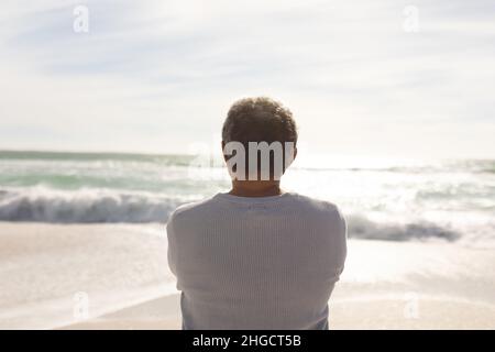 Vista posteriore dell'anziano biraciale che guarda onde che si infrangono in mare contro il cielo dalla spiaggia soleggiata Foto Stock