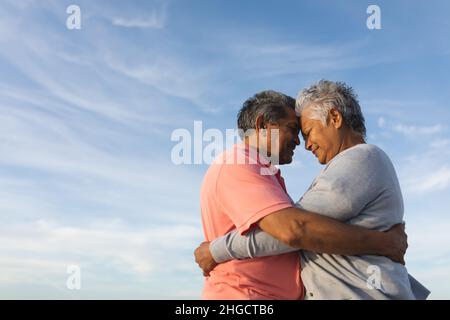 Vista laterale della coppia anziana multirazziale affettuosa che abbraccia mentre tocca la fronte contro il cielo Foto Stock