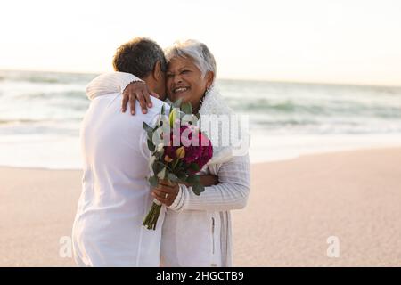 Allegro newlywed coppia anziana multirazziale che tiene il bouquet del fiore che abbraccia alla spiaggia durante il tramonto Foto Stock