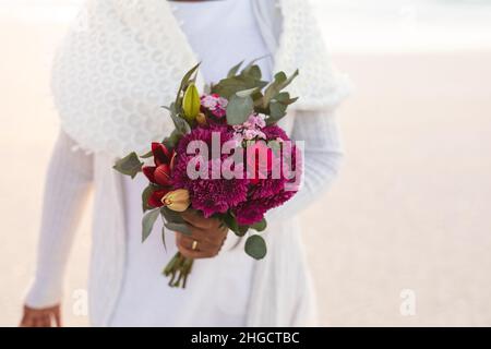 Sezione centrale di donna biraciale senior che tiene bouquet di fiori freschi in spiaggia durante il tramonto Foto Stock