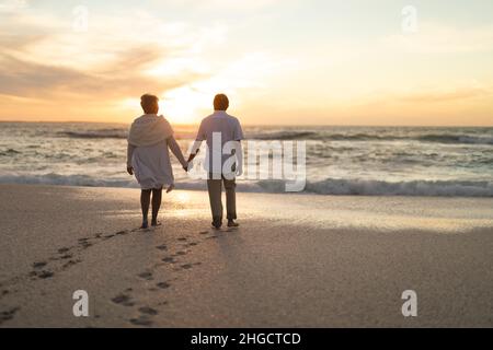 Vista posteriore della coppia anziana multirazziale newlywed che tiene le mani che camminano alla spiaggia durante il tramonto Foto Stock