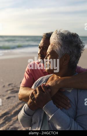 Uomo biraciale anziano e premuroso che abbraccia la donna da dietro mentre guarda insieme in spiaggia Foto Stock
