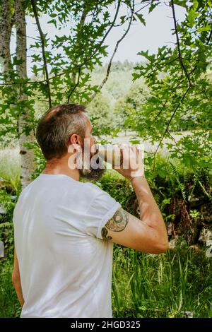 Uomo tatuato che beve un sano frullato all'esterno circondato da verde paesaggio naturale in campagna Foto Stock