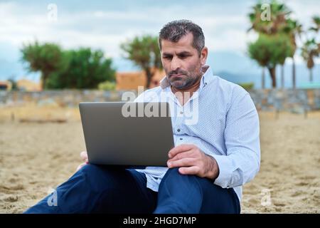 Grave uomo maturo triste con un computer portatile seduto su una spiaggia nuvolosa. Foto Stock
