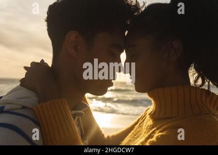 Vista laterale di coppia multirazziale romanticismo mentre si abbracciano a vicenda in spiaggia durante il tramonto Foto Stock