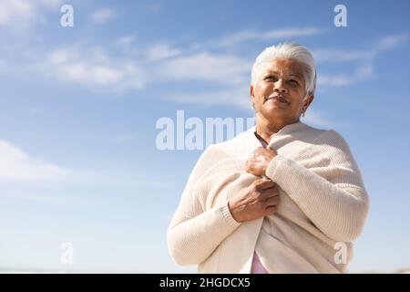 Vista ad angolo basso della donna anziana biraciale che indossa un tappeto guardando via alla spiaggia contro il cielo blu Foto Stock