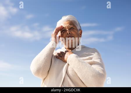 Vista ad angolo basso della donna anziana biraciale che scherma gli occhi mentre guarda via contro il cielo nelle giornate di sole Foto Stock