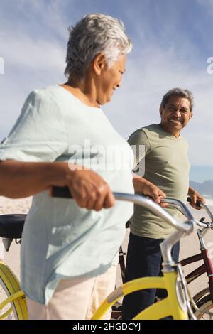Felice coppia multirazziale senior che si guardano l'un l'altro mentre si sta in piedi con le biciclette sulla spiaggia soleggiata Foto Stock