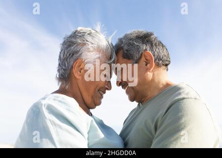 Vista laterale di sorridente coppia multirazziale senior che tocca la fronte in spiaggia durante la giornata di sole Foto Stock