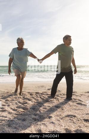 Tutta la lunghezza dell'uomo biraciale anziano che tiene la mano con la donna mentre la conduce alla spiaggia contro il cielo Foto Stock