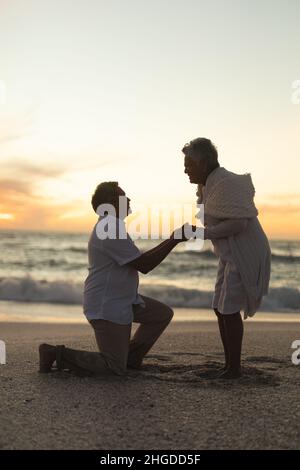 Vista laterale dell'uomo biraciale anziano che propone la donna mentre si inginocchiava sulla spiaggia durante il tramonto Foto Stock