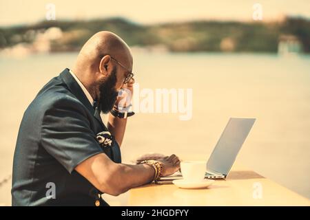 Un serio imprenditore africano-americano concentrato uomo sta lavorando sul suo laptop in un caffè all'aperto accanto al fiume; un elegante calvo bearded nero Foto Stock