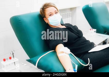 Donna caucasica di mezza età dona sangue da vena in ospedale. Il donatore si siede in sedia medica che indossa la maschera medica e guarda direttamente nella cellula. Scena reale. Persone reali. Aiutare covid 19 pazienti Foto Stock