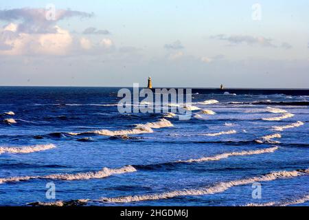 Onde che si avvolgono sulla riva a Cullercoats, Inghilterra, con il faro sul molo di Tynemouth sullo sfondo Foto Stock