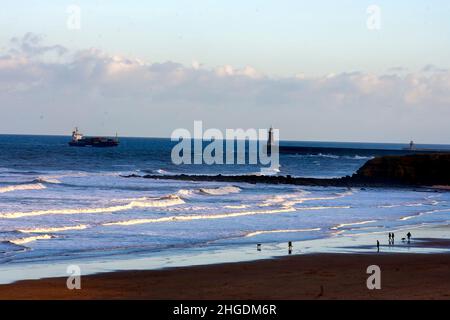 Onde che si avvolgono sulla riva a Cullercoats, Inghilterra, con il faro sul molo di Tynemouth sullo sfondo Foto Stock