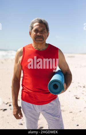 Ritratto di fidato fit biraciale uomo anziano che tiene arrotolato tappeto yoga sulla spiaggia contro il cielo blu Foto Stock