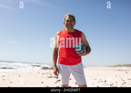 Ritratto di sorridente uomo biraciale anziano che tiene laminato yoga mat a spiaggia soleggiata contro il cielo blu Foto Stock