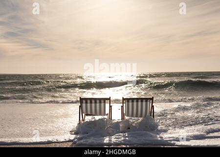 Onda che spruzzi su due sedie di legno pieghevoli vuote in spiaggia contro il cielo durante il tramonto Foto Stock