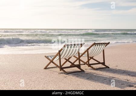 Due sedie di legno pieghevoli e vuote sulla spiaggia di fronte alle onde che si infrangono in mare contro il cielo Foto Stock