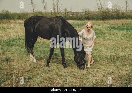 Una bella giovane donna in un abito in chiffon bianco con un breve taglio di capelli biondo e gioca con un cavallo nero in un prato. Foto Stock