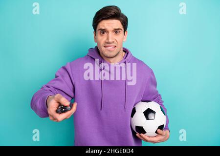 Foto di giovane ragazzo nervoso turbato vestito felpa viola tenendo palla di calcio cambiare canali tv isolato sfondo di colore turchese Foto Stock
