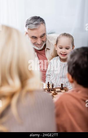 uomo anziano sorridendo mentre giocava a scacchi con nipoti e moglie, offuscato primo piano Foto Stock