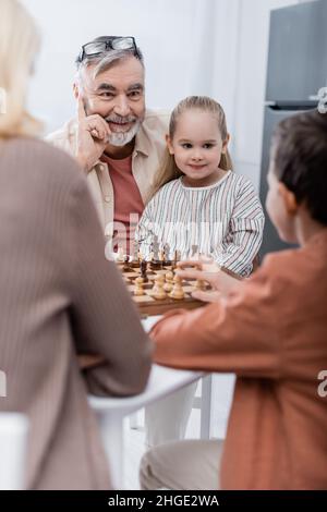 ragazzo allegro che gioca a scacchi con moglie e nipoti in primo piano sfocato Foto Stock