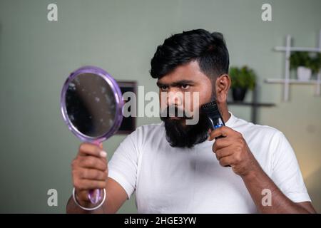 Giovane uomo indiano della barba che usa il rasoio elettrico o la macchina di trimbere guardando il concetto specchio- della cura dei capelli, del grooming e del wellness. Foto Stock