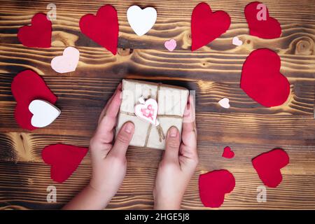 Mani che tengono una scatola in carta kraft con un arredamento a forma di cuore su uno sfondo decorato per San Valentino. Regalo per un concetto amato uno. Messa a fuoco soft Foto Stock