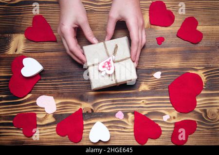 Mani che tengono una scatola in carta kraft con un arredamento a forma di cuore su uno sfondo decorato per San Valentino. Regalo per un concetto amato uno. Messa a fuoco soft Foto Stock