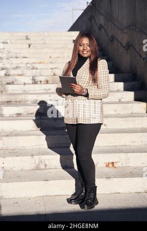 Donna d'affari afroamericana che usa un tablet. Foto verticale con donna sorridente su una scala. Foto Stock