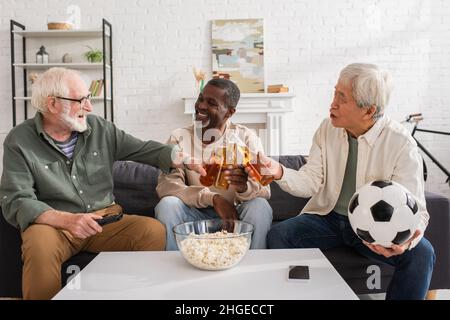 Amici multiculturali che scannano birra vicino al calcio e popcorn a casa Foto Stock