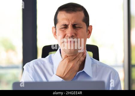 Scatto di un giovane uomo d'affari che avverte dolore alla gola mentre lavora in un ufficio Foto Stock