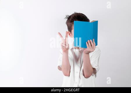 La mano del ragazzo piccolo che mostra la vittoria, leggendo un libro e coprendo il suo volto su sfondo bianco. Foto Stock