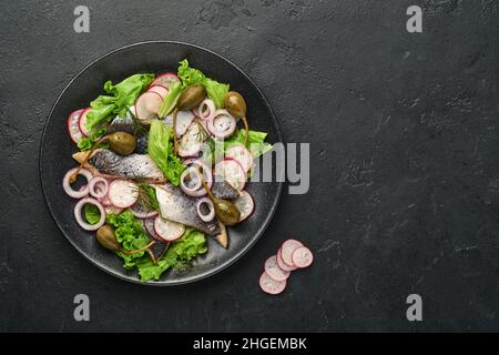 Aringhe salate con spezie, capperi, erbe e insalata di cipolla su piatto nero su sfondo scuro con spazio copia. Pesce marinato a fette. Cibo con u sano Foto Stock