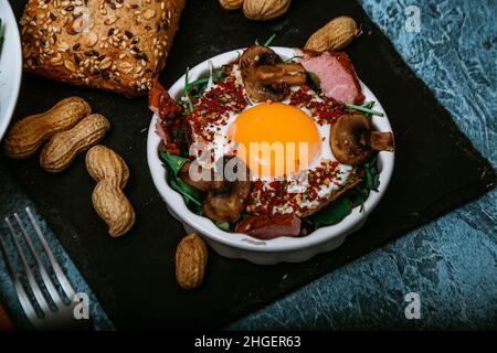 Uova fritte cucinate perfettamente con funghi, spezie, pepe, carne e sale su rucola. Piatto colazione rotondo su tavola nera con arachidi e baguette w Foto Stock