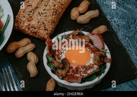 Uova fritte cucinate perfettamente con funghi, spezie, pepe, carne e sale su rucola. Piatto colazione rotondo su tavola nera con arachidi e baguette w Foto Stock