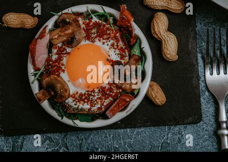 Uova fritte cucinate perfettamente con funghi, spezie, pepe, carne e sale su rucola. Piatto colazione rotondo su tavola nera con arachidi e baguette w Foto Stock