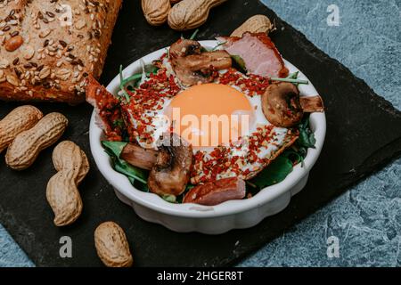 Uova fritte cucinate perfettamente con funghi, spezie, pepe, carne e sale su rucola. Piatto colazione rotondo su tavola nera con arachidi e baguette w Foto Stock