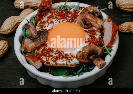 Uova fritte cucinate perfettamente con funghi, spezie, pepe, carne e sale su rucola. Piatto colazione rotondo su tavola nera con arachidi Foto Stock