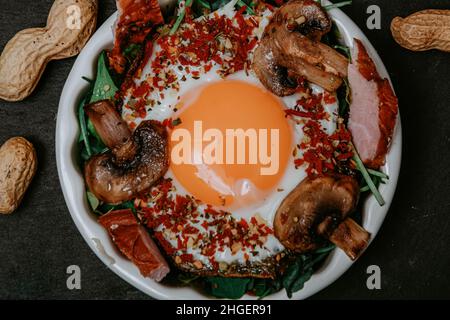 Uova fritte cucinate perfettamente con funghi, spezie, pepe, carne e sale su rucola. Piatto colazione rotondo su tavola nera con arachidi Foto Stock