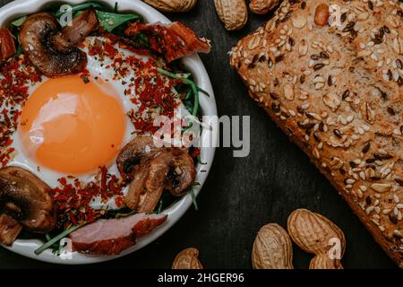 Uova fritte cucinate perfettamente con funghi, spezie, pepe, carne e sale su rucola. Piatto colazione rotondo su tavola nera con arachidi e baguette w Foto Stock