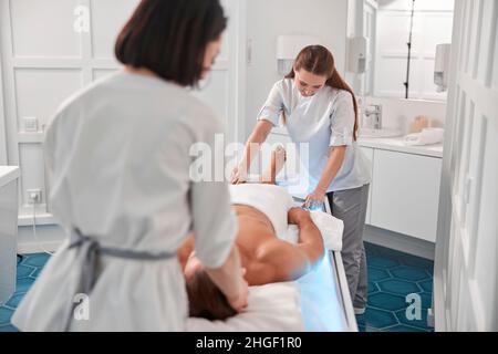 I terapisti lavorano con le gambe dell'uomo utilizzando uno strumento speciale, mentre i colleghi massaggiano la testa in ospedale Foto Stock