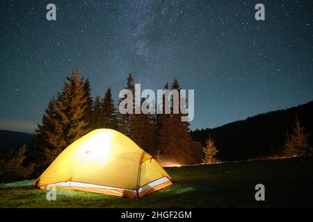 Luminosa tenda turistica illuminata che risplenderà sul campeggio in montagne scure sotto il cielo notturno con stelle scintillanti. Concetto di stile di vita attivo. Foto Stock