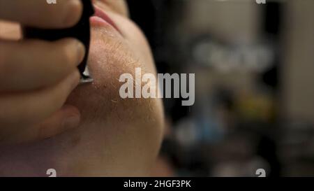Primo piano ritratto della vista laterale del giovane uomo bearded che ottiene la barba grooming nel barbiere moderno in slow motion. Parrucchiere al servizio del cliente, fare la barba Foto Stock