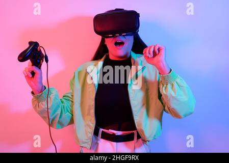 Una ragazza spaventata al neon con occhiali VR tiene in mano un gamepad e gioca. Mondo virtuale, tecnologia. Emozioni di paura, orrore, horror ga Foto Stock