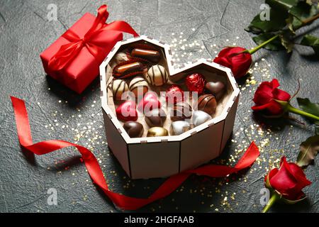 Scatola a forma di cuore con gustose caramelle al cioccolato, regalo e rose su sfondo nero Foto Stock
