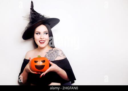 Una ragazza vestita come una strega sta tenendo una zucca. Modello in cappello con tatuaggio sulla spalla sorridente Foto Stock