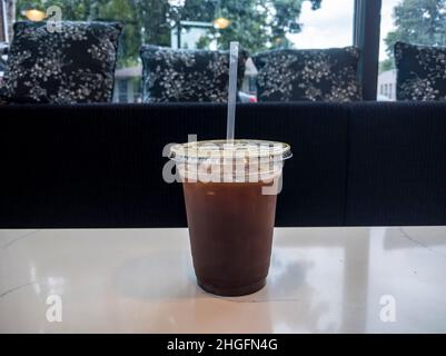 Caffè freddo e ghiacciato in una tazza di plastica con cannuccia, seduto su un tavolo bianco in un accogliente caffè con panca imbottita nera Foto Stock